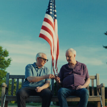 Two older men wearing meta glasses sitting on a bench with american flag behind them.